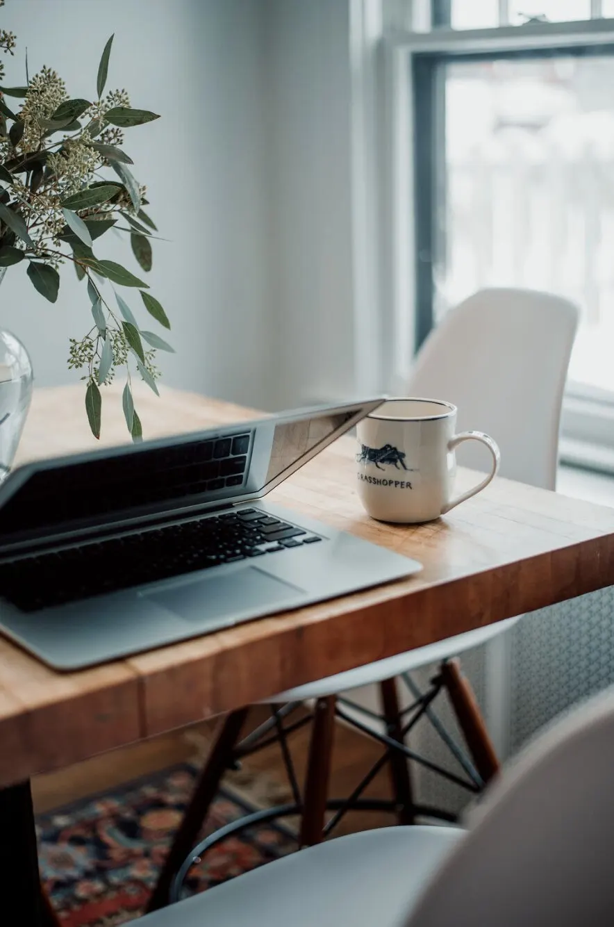 A laptop and a white Grasshopper mug on a butcher-block table by a bright window, with a sprig of eucalyptus in a glass vase.
