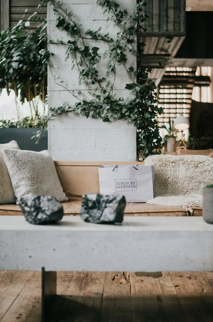 A sunlit lounge with ivy climbing a white brick pillar, a tan sofa with sheepskin cushions, a Bergdorf Goodman shopping bag, and two dark stones on a concrete table.