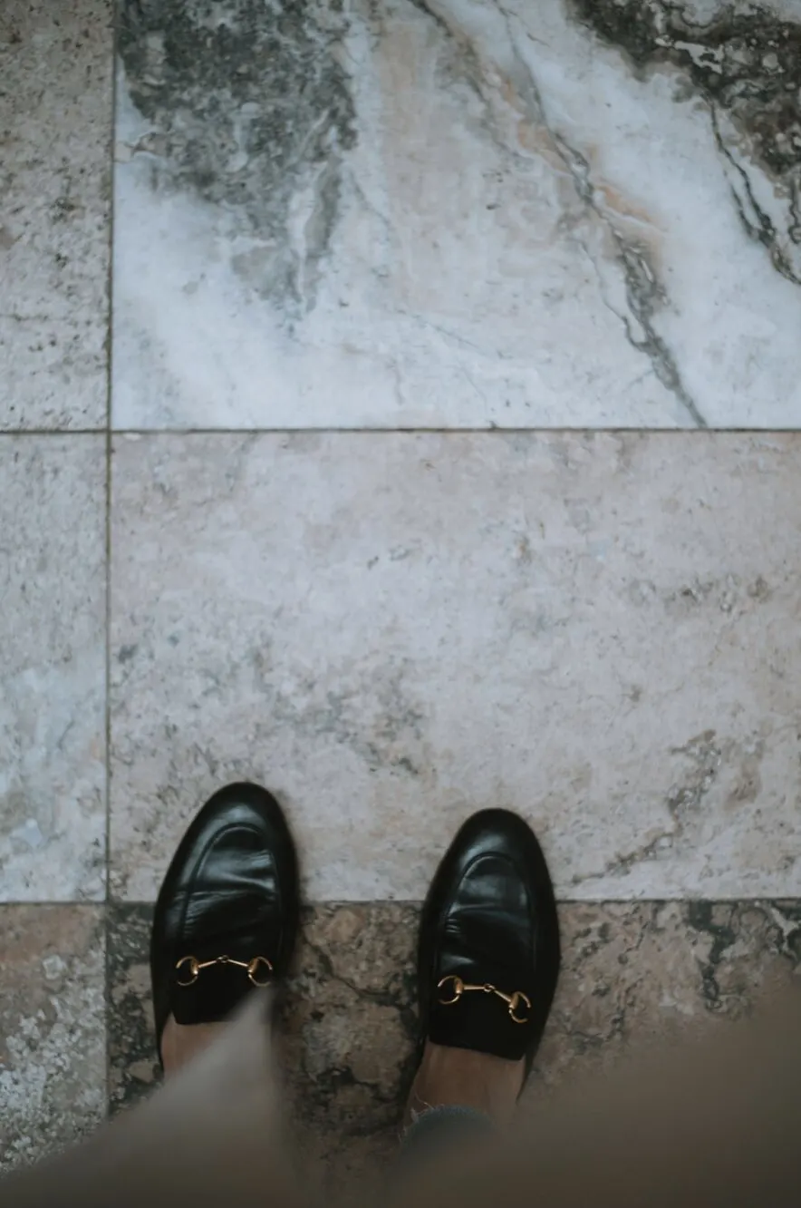 Looking down at a pair of black leather horsebit loafers standing on pale marble floor tiles.