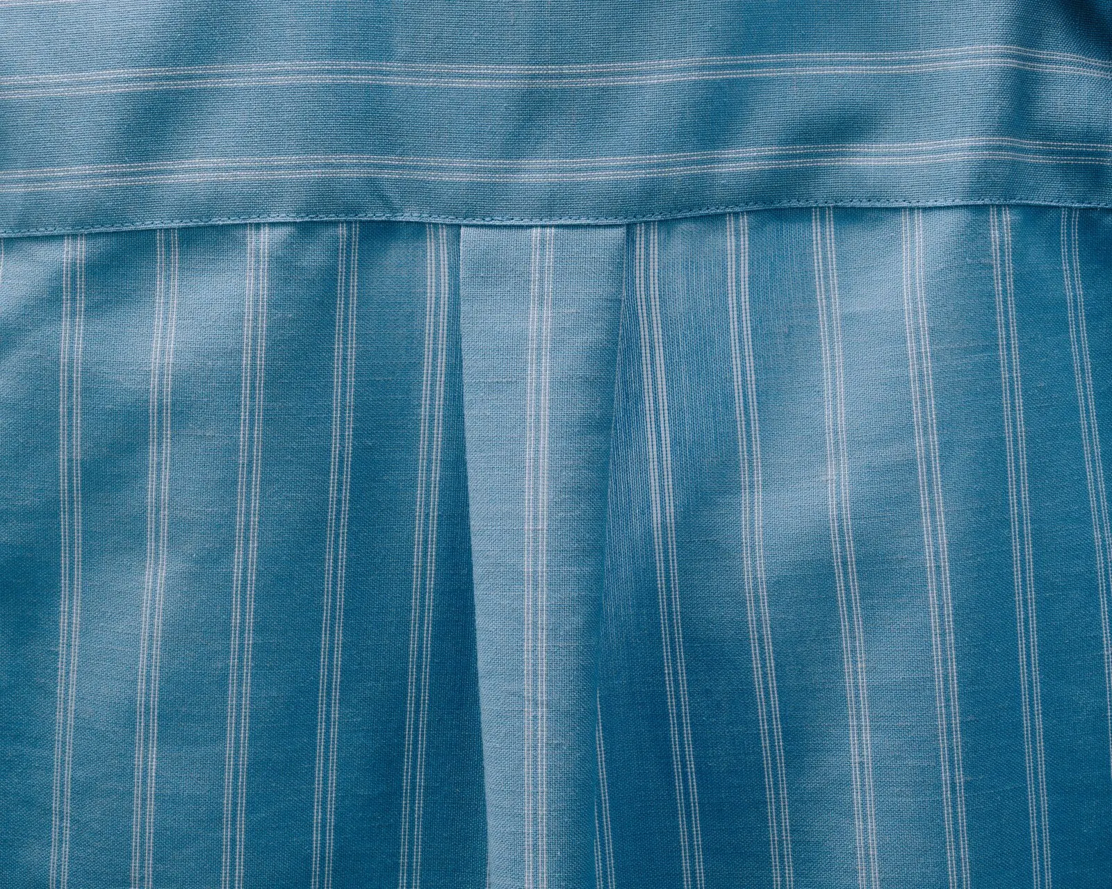 A close-up of blue pinstriped shirting at the back yoke, where a stitched seam meets a soft box pleat.