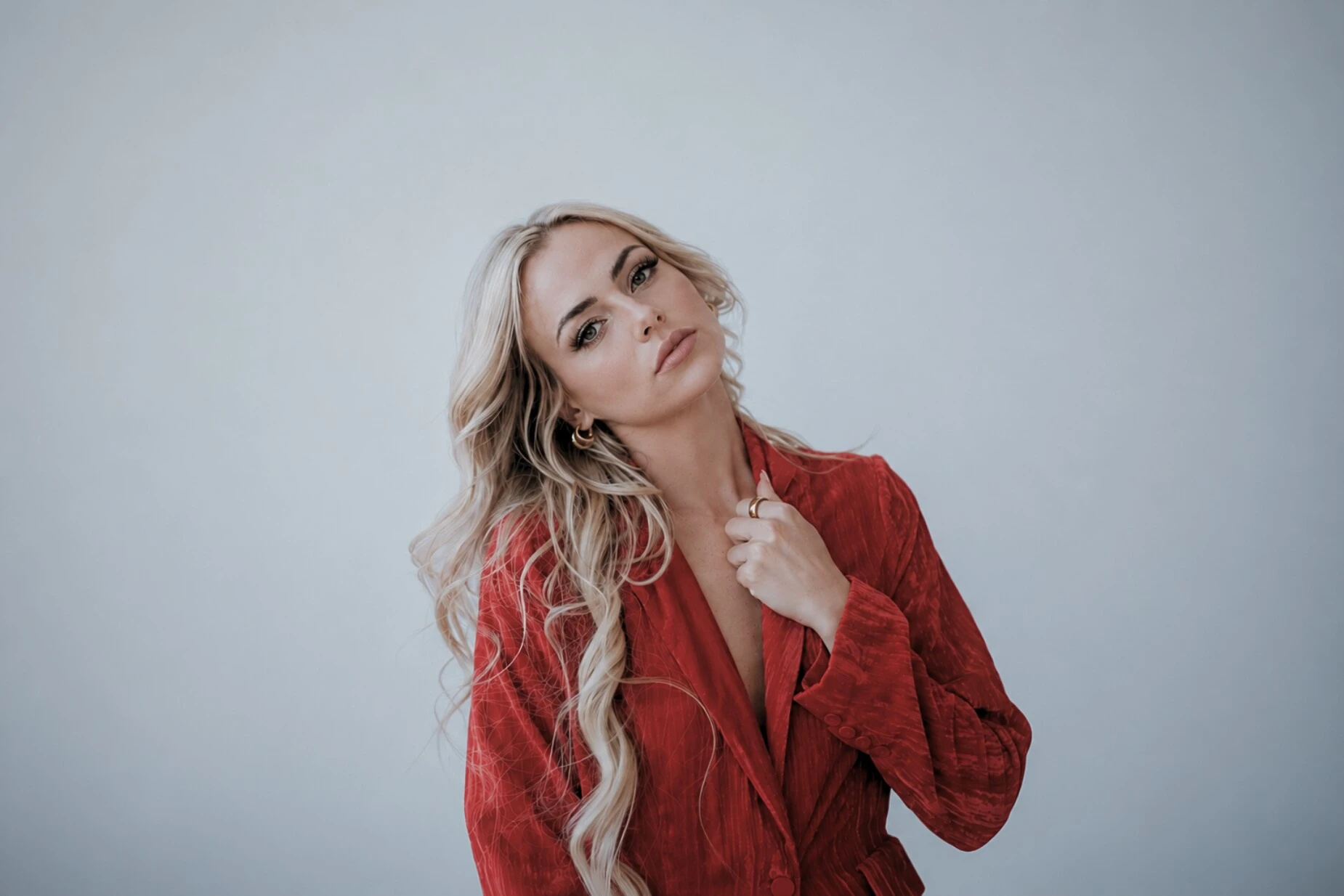Tyler Tanner in a crimson crushed-velvet blazer against a pale grey studio wall, head tilted, one hand at her collar, a gold ring and gold hoops.
