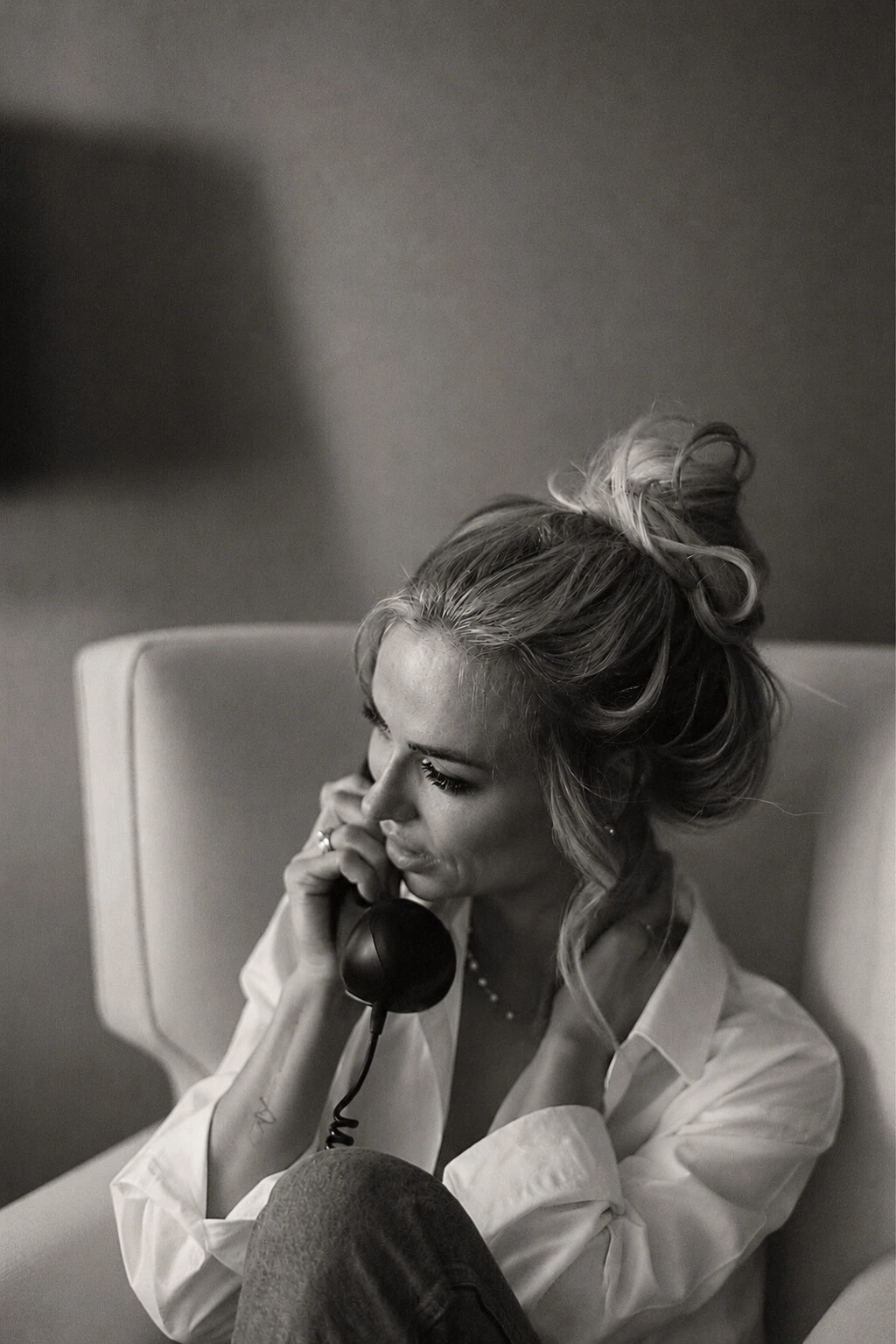 Black and white portrait of a woman with her hair loosely pinned up, in a white shirt, seated in a pale armchair speaking on a black telephone receiver.