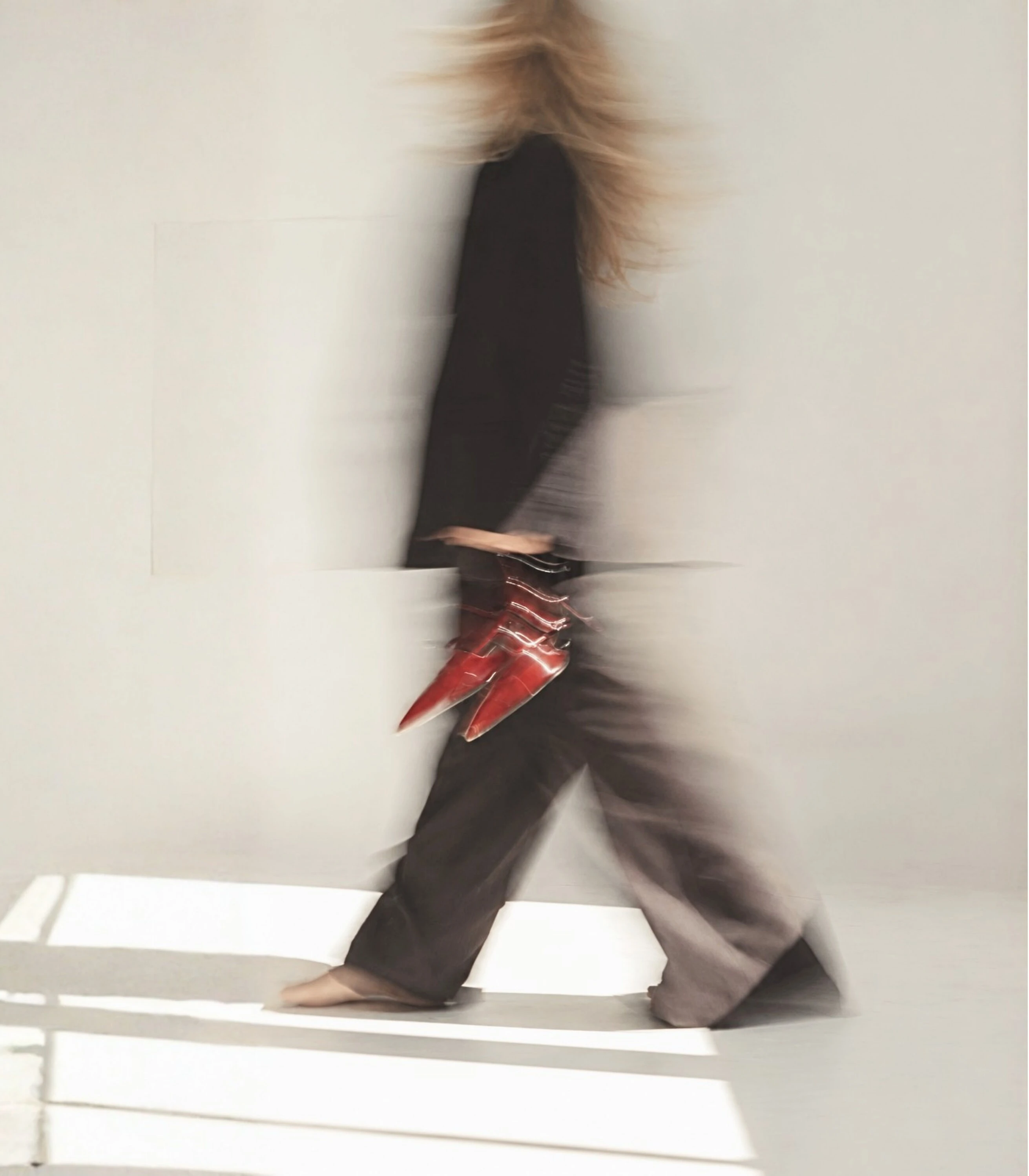 A woman in a black top and wide brown trousers walking barefoot across a pale studio in a blur of motion, red patent shoes in hand.