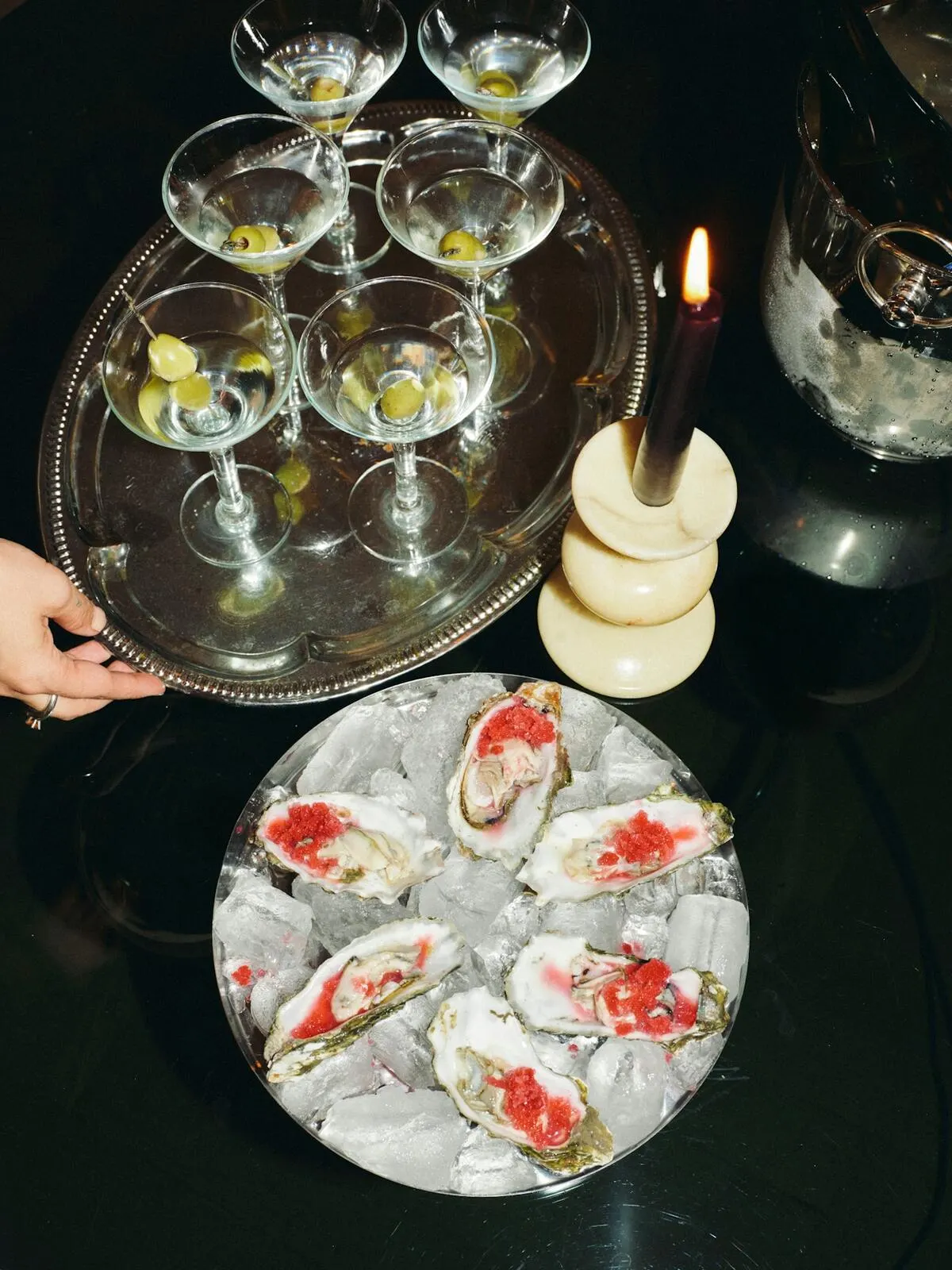 A silver tray of six martinis and a plate of oysters dressed with roe on ice, a lit candle and a champagne bucket on a black table.