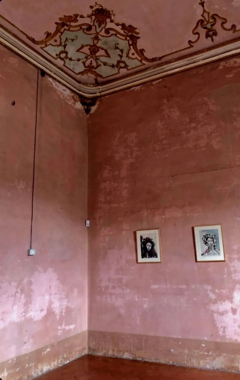 An empty room with distressed pink plaster walls, ornate cornice, and two small framed pictures.