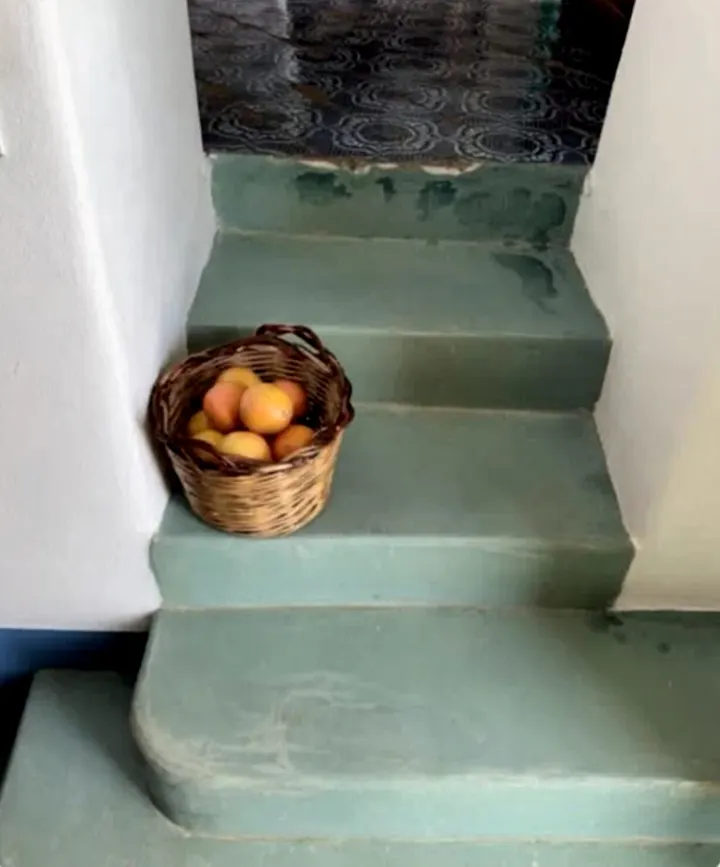 A wicker basket of apricots left on worn sea-green stone steps beside a whitewashed wall.