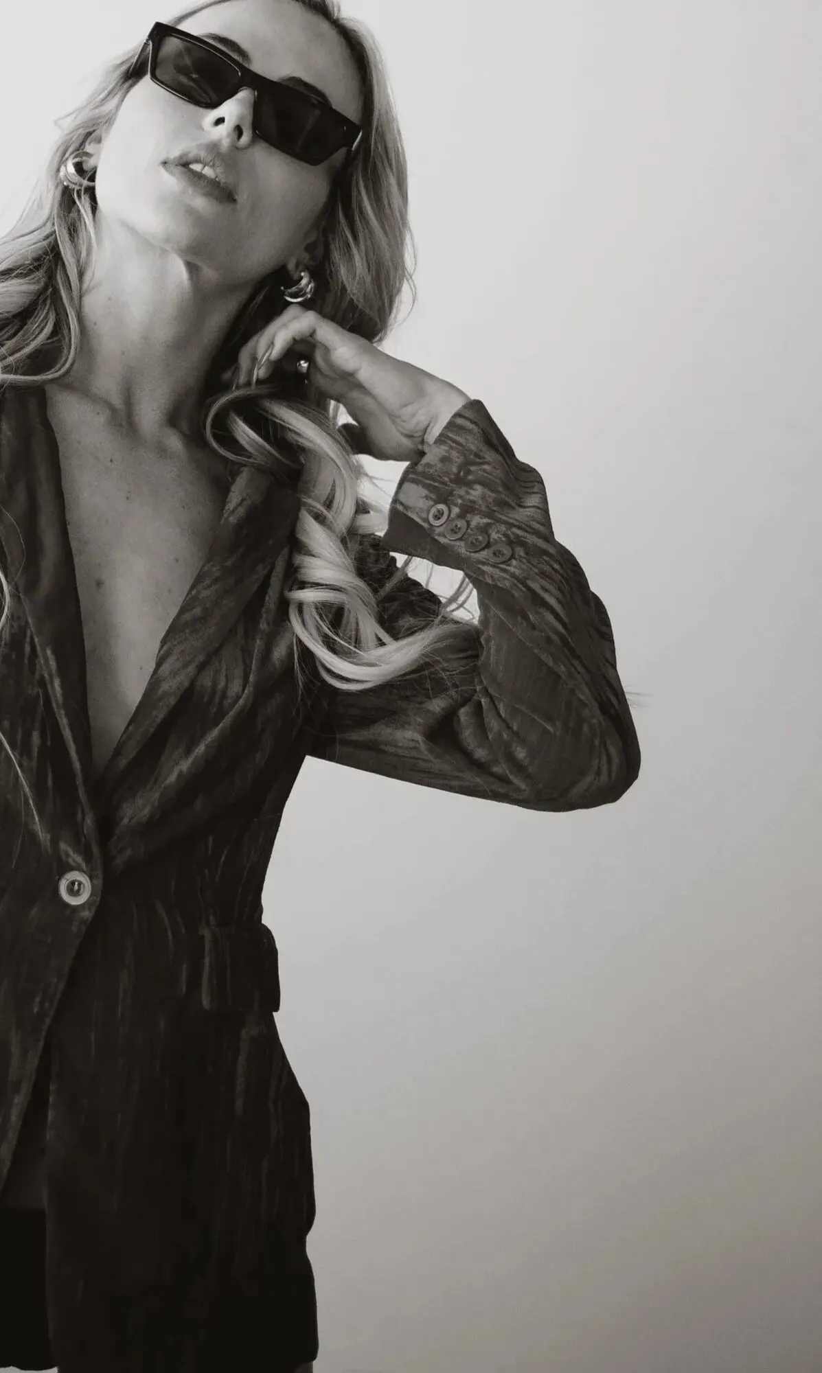 Tyler Tanner in an open velvet blazer and dark sunglasses against a pale studio wall, black and white.