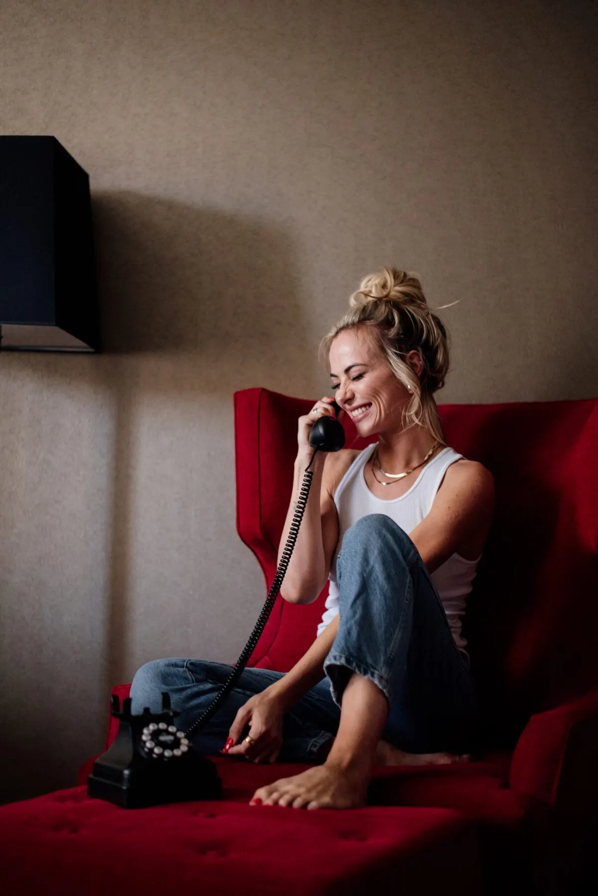 Tyler Tanner sitting barefoot on a red velvet chaise in a white tank and jeans, laughing into the handset of a black rotary telephone.