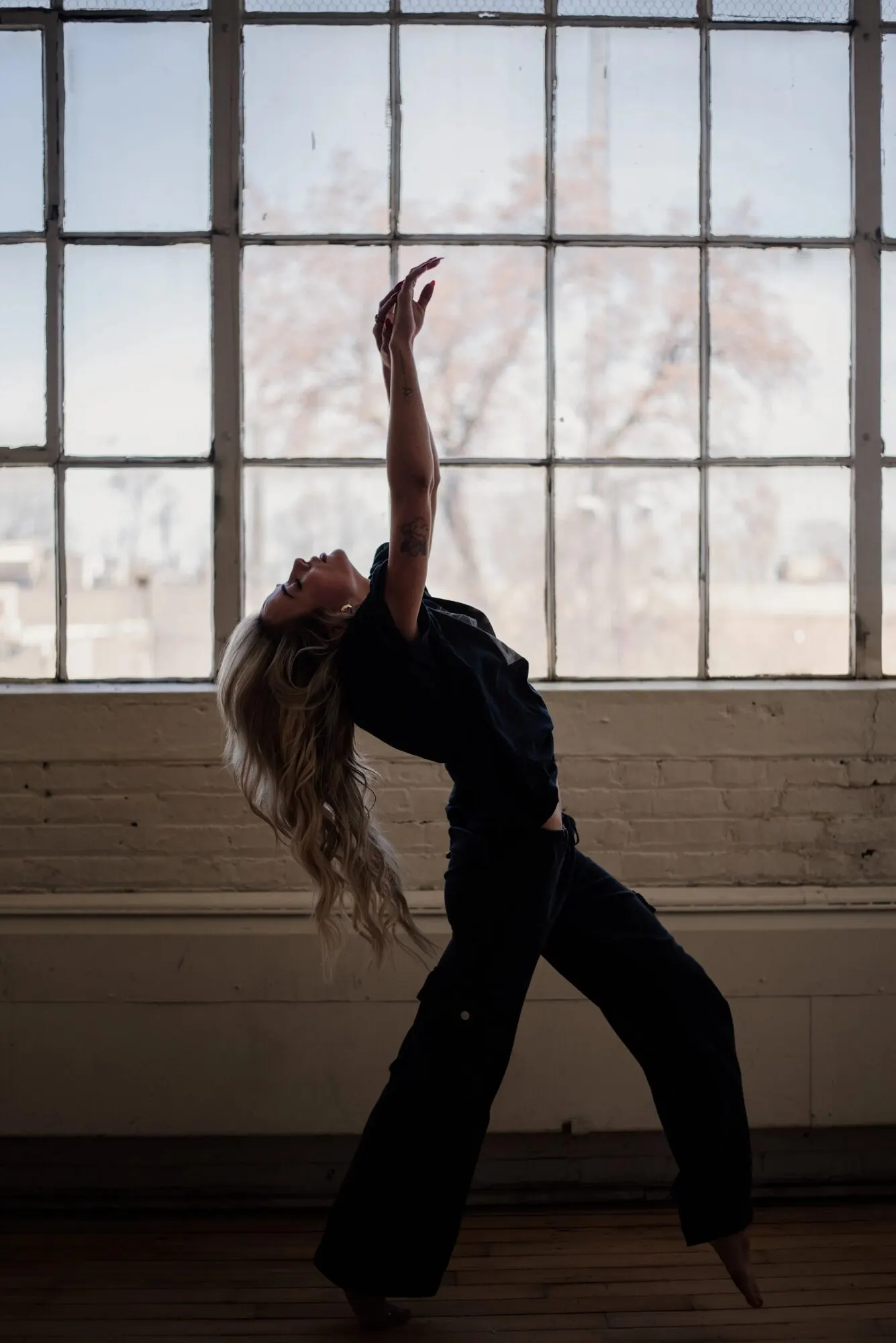 Figure caught mid-movement, back arched, against a tall grid of factory windows.