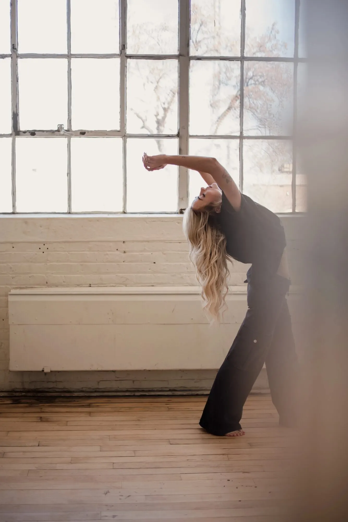 Tyler Tanner arched back mid-stretch in front of a wall of factory windows, barefoot on a wood floor.